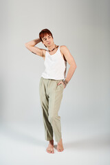 A stylish young man striking a pose in a white tank top and khaki pants against a grey studio background.
