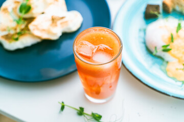 Poached eggs with hollandaise sauce on a turquoise plate with grapefruit juice, cutlery, cheese and avocado on a white table. Selected focus