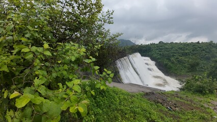 A dam with water flowing, surrounded by lush greenery under a cloudy sky, showcasing the beauty of...