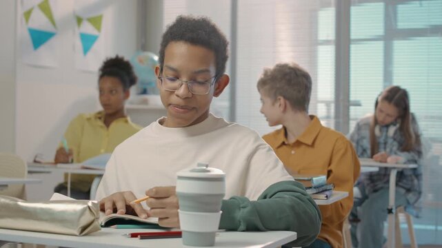 Medium shot of African American teenage boy sitting at desk doing tricks with pencil and fooling around among classmates laughing during lesson at school