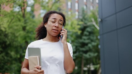 Portrait of talkative woman strolling near contemporary building during communication process on personal smartphone. Young female dressed in casual white t-shirt holding portable laptop outdoors.