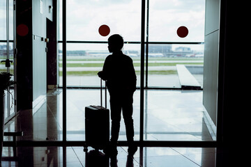 silhouette of caucasian boy with suitcase standing by the window in airport watching planes waiting for flight. Travelling concept
