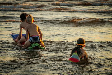 Three children playing in the water at the beach during sunset. The girl in a colorful swimsuit...