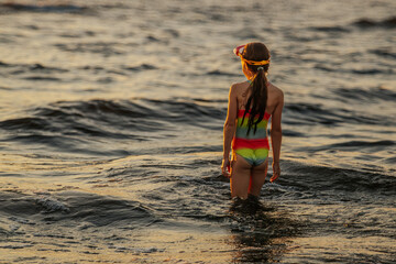 A girl in a colorful swimsuit and pink goggles playfully splashing in the waves at the beach during...