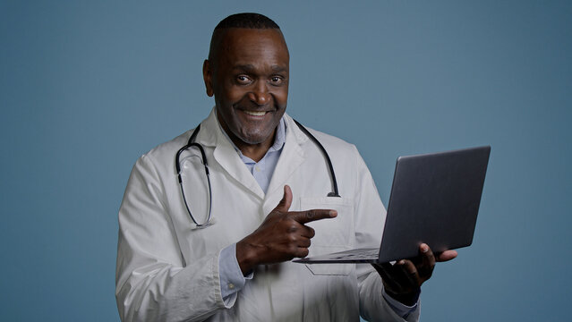 African american man doctor therapist ethnic male surgeon in white coat in blue background studio working on laptop consulting patient online remote diagnostics take useful information from internet