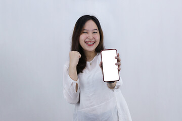 Cheerful young Asian woman holding smart phone with fists clenched celebrating victory expressing success on white background.