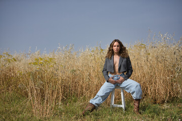 Young woman, beautiful, blonde, with green eyes, jacket, jeans and cowboy boots, sitting on stool, feminine, free, independent and sensual. Concept beauty, countryside, freedom, sensuality, nature.