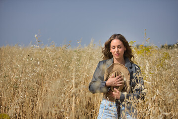 Young woman, farmer, beautiful, blonde, with closed eyes, jacket and jeans, covering her chest with a hat, in the middle of an oat field, sad and melancholic. Concept beauty, field, melancholy, nature