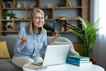 Smiling senior woman recording podcast, sitting on sofa with laptop and microphone. Engaged in...
