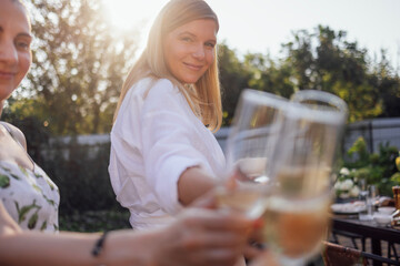 Young woman with glass of wine making toast outdoors. Attractive blonde clinks goblet of wine with her friends in backyard of house.