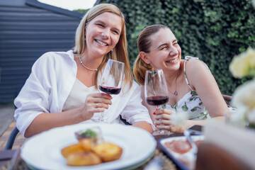 Two young women in white dresses are having lunch outside. Charming friends make a toast and clink glasses on the terrace.