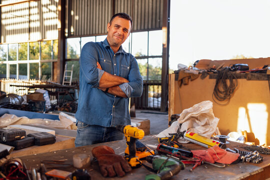 A young smiling man in casual clothes stands at a table with construction tools. An attractive builder at a warehouse workplace.