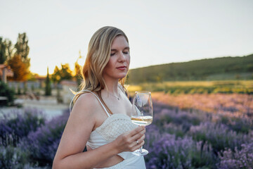Young female blonde in a white dress and a straw hat holds a glass of wine in a lavender field