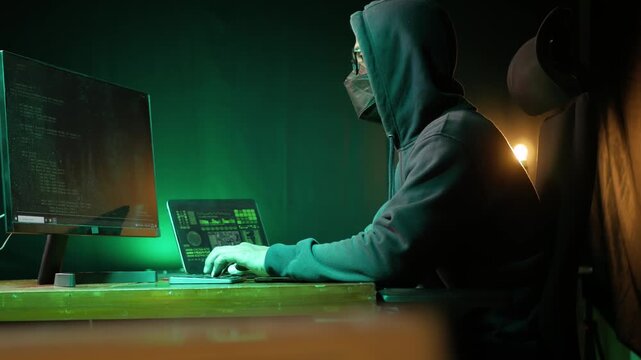 Hooded asian male hacker wearing a black mask sitting at dark room in front of pc monitor, writing code or using computer virus program for cyber attack. cybercrime, hacking concept