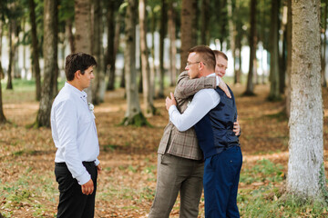 Three men hug each other in a forest. One of them is wearing a vest. Scene is warm and friendly