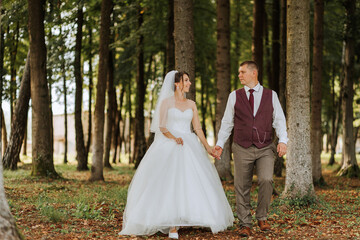 A bride and groom are walking through a forest, hand in hand. The bride is wearing a white dress and the groom is wearing a vest. Scene is romantic and peaceful