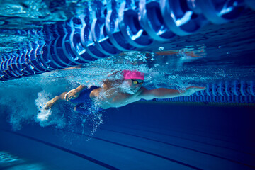 Competitive swimmer, with pink cap and blue trunks, seen underwater during dive, demonstrates his...