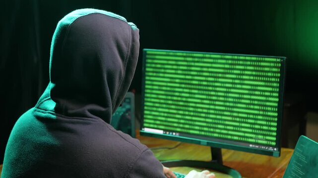 Back view of hooded computer hacker sitting at dark room in front of pc monitor, writing code or using computer virus program for cyber attack. cybercrime, hacking concept