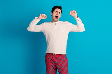 Portrait of nice young man yawn stretch arms wear pajama isolated on blue color background