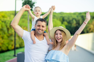 happy young parents mom and dad are walking with a baby in a stroller in the park in a hat in the summer in the setting sun and smiling having fun