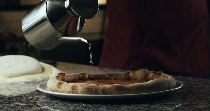 Super slow motion close up of pizzaman puts fresh olive oil on traditional italian dish of pizza margherita with tomato sauce and mozzarella cheese before save it on restaurant kitchen at 1000 fps.