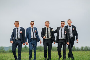 A group of five men are dressed in suits and ties, walking together in a field. Scene is one of...
