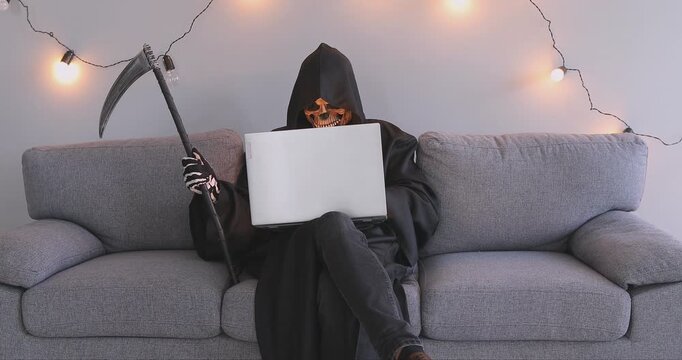 Halloween Death man with laptop online, bad horrible hacker looking for new victim, Grim Reaper cosplay costume with scythe, scary man black hooded cape, skull face mask, skeleton gloves party spooky