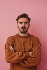 Obraz premium Man standing with arms crossed, against a pink wall. He has a beard and is wearing brown sweater.