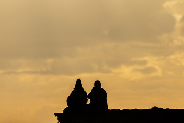 Silhouette of the backs of two people enjoying the sunset sitting on a rock looking out to sea