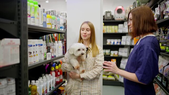 A blonde girl with a white dog listens to a pet store assistant in a blue uniform and makes a decision about purchasing goods in the pet store