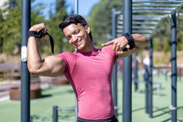 Smiling man in a pink shirt flexing biceps and pointing in an outdoor gym
