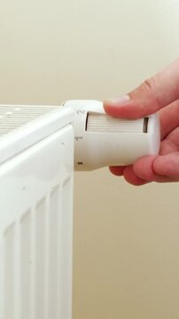 Unrecognizable man's hand setting temperature on central heating system. White metal heating iron household radiator on white wall inside room. Thermostat at home near white wall. Budget in crisis