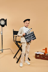 A preadolescent boy in film director attire holds a movie clapper in front of a camera.