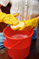 Woman in yellow gloves cleaning red bucket with sponge on wooden table in front of another woman in...