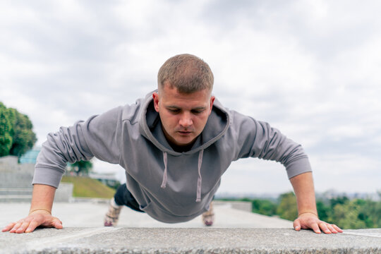 in the park, a young guy in a gray hoodie is doing a warm-up doing push-ups on the camera healthy lifestyle cloudy weather - Powered by Adobe