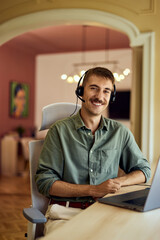 Fototapeta premium Smiling man working at the office, wearing headphones, using a laptop.