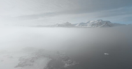 Drone fly foggy sky over Antarctica ocean, snow-capped mountain peaks, sunrise clouds in background. Morning mist over Antarctic coast, fog cloudscape. Aerial view surface. Wild nature background