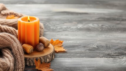 An orange candle burns on a wooden slab surrounded by autumn leaves, acorns, and a brown knit blanket