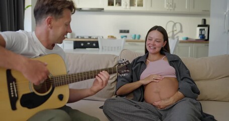 Happy pregnant brunette woman sitting on the sofa and listening to her husband playing guitar at home on the weekend - Powered by Adobe