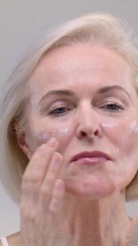 Portrait: old woman in the studio used white anti age product on the cheek. Beautiful elderly female puts lotion on her face with her hand. Senior lady uses a moisturizing cream for the skin of head