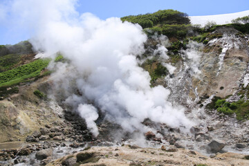 Geothermal White Steam Vents releasing plumes in Rocky Terrain surrounded by lush green vegetation and patches snow under bright blue sky. Natural phenomenon dynamic interaction