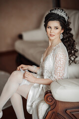 A woman in a white gown sits on a chair with her legs crossed. She is wearing a tiara and has her...