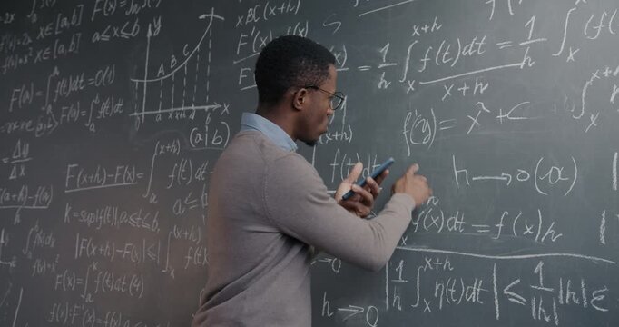 Science teacher African American man checking formula on blackboard and using smartphone in university classroom. Education and modern technology concept.
