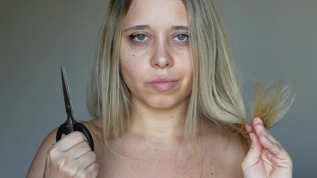 A young caucasian blonde woman with dry bleached hair holding scissors in her hands going to cut the ends of her hair on a grey background. Hair care