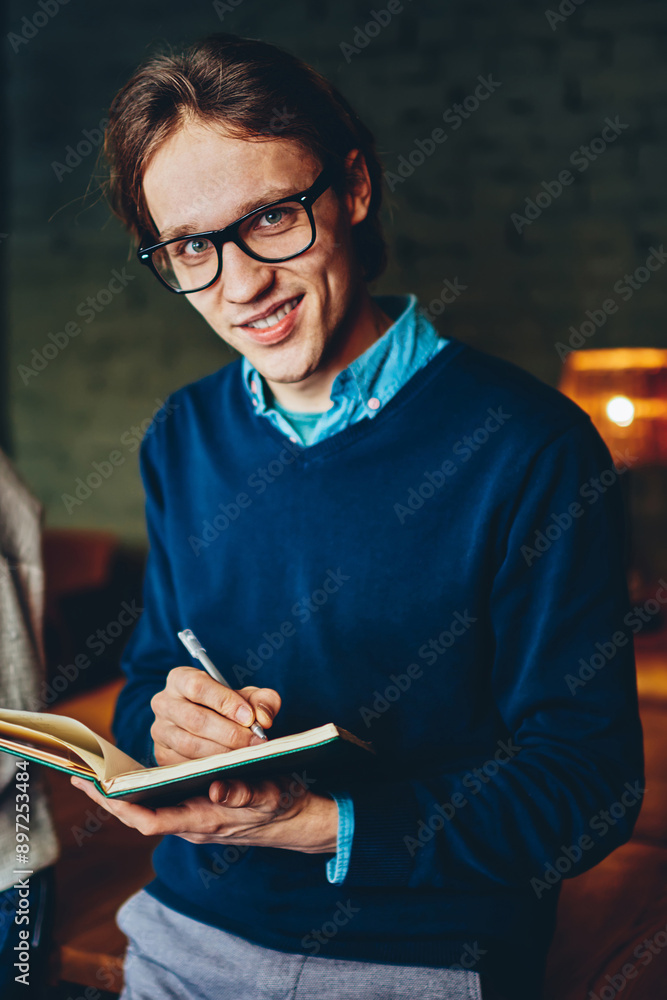 Poster skilled male in eyewear making notes in notepad looking at camera during working process in office w
