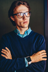Pensive young male entrepreneur in spectacles for vision correction thinking about idea for startup project looking away,thoughtful businessman with crossed arms and smartwatch standing indoors