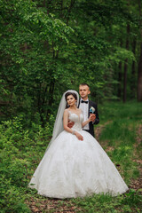 A bride and groom are posing for a picture in a forest. The bride is wearing a white dress and the groom is wearing a black suit. They are holding hands and smiling for the camera