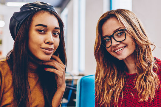 Portrait of successful multicultural hipster girls looking at camera and enjoying coffee break indoors, happy smiling female teenagers in trendy apparel holding takeaway cup at campus on leisure