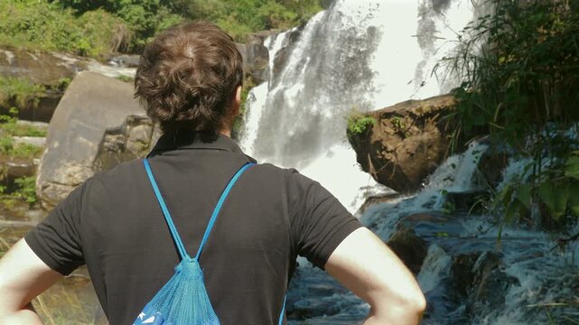 Rear view of person standing with hands on hips, wearing blue drawstring backpack, admiring beautiful waterfall in lush, green, forested area. Adventure and Nature.