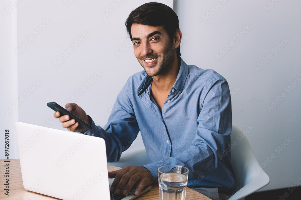 Poster smiling successful male graphic designer working with laptop device in modern workspace, positive ma